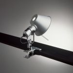 Artemide Tolomeo clip task lamp in polished aluminum, conical shade head mounted directly on a metal spring clip clamped to a black bar, three quarter view.